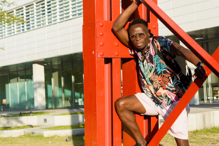 young fashionable african man, posing in summer clothes, leaning against some orange beams, looking at the camera.の写真素材