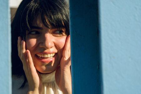 portrait of young latin woman, surprised smiling with open mouth, looking to side and hands on face, over blue beams, people concept, copy space.の写真素材