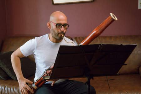 front view of young latino man of venezuelan ethnicity, bald with glasses and white clothes, classical music student sitting at home, practicing with bassoon, reading sheet music on music stand.の写真素材
