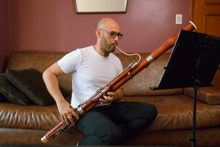 young latino man, bald with glasses and a white shirt, is sitting on a leather sofa playing the bassoon, reading and studying sheet music at home.の写真素材