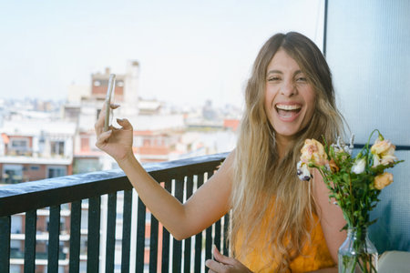 young latin woman of colombian ethnicity sitting on the balcony of her apartment holding her phone...