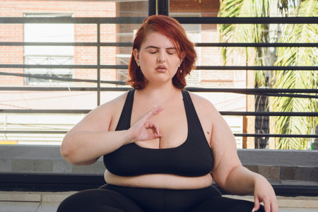 young latin plus size woman sitting with eyes closed meditating with hand on chest in gyan mudra position, front view indoors, yoga concept.の写真素材