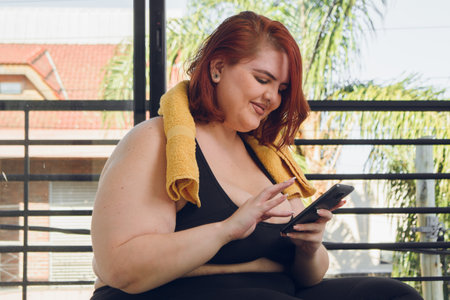 young plus size latin woman of argentinian ethnicity in sportswear sitting indoors smiling and using her phone touching the screen, healthy life concept, copy space.の写真素材
