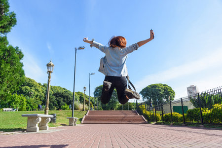 rear view of young woman jumping in the park with her legs and arms open, she is dressed in a jean...