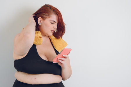worried young plus size woman reading messages on her phone holding her head, wears black sportswear and short red hair, standing over white background, copy spaceの写真素材