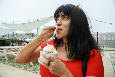 young happy latin woman outdoors eating ice cream, holding a small cup with mixed flavors. copy...