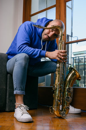 vertical portrait of young venezuelan latino man, at home sitting on the sofa in the living room with his saxophone calm and content looking at the camera.の写真素材