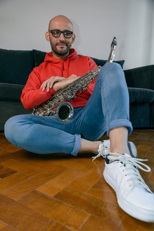 vertical portrait of young latin musician man with beard, glasses and red sweater with blue pants, sitting on the floor with his saxophone happily looking at the camera.の写真素材