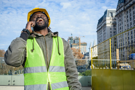 African ethnicity engineer man with a beard, wears a yellow safety helmet and reflective vest, very happy smiling out loud outdoors talking on the phone, copy space.の写真素材