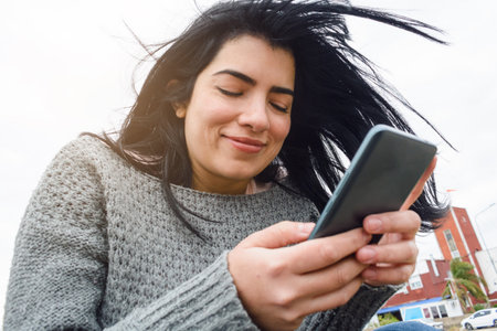 young venezuelan latina woman, using the phone, reading messages, browsing the internet, calmly sitting outdoors on a cloudy and cold day, dressed in gray and with her hair moved by the wind.の写真素材