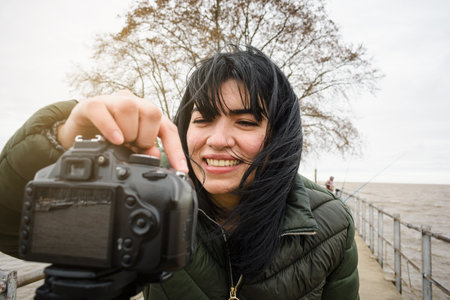closeup young latin woman vlogger happy smiling pressing the record button on her digital DSRL camera to make a video standing on the pierの写真素材