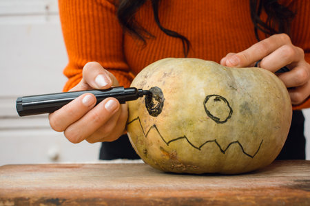 front view closeup of female hands of unrecognizable person, drawing with black marker eye on pumpkin on wooden board, decorating for halloween night, copy space, crafts concept.の写真素材