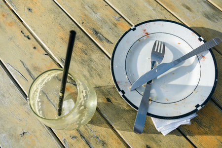 top view of dirty porcelain plate with recently used fork and teaspoon on table next to empty glass with recently used straw, outside restaurant at dusk. copy space.の写真素材