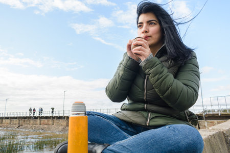 young latin woman sitting outdoors on cold day, sitting enjoying morning drinking yerba mate,...