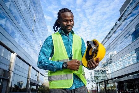 young civil engineer man of african ethnicity with beard and dreadlocks, holding his helmet with hand is standing outdoors smiling happy at end of work dayの写真素材