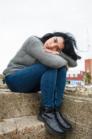 young melancholic latin woman sitting sad pensive and calm with crossed arms leaning on her legs outdoors on cold and cloudy day, people concept, copy spaceの写真素材