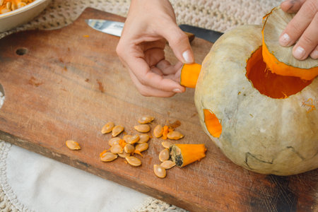 closeup of unrecognizable female caucasian hands removing portion to make eye of pumpkin on wooden cutting board, decorating it for Halloweenの写真素材