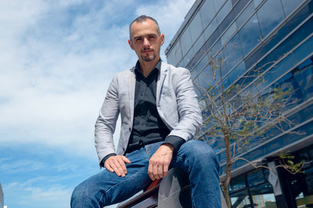 portrait of elegant caucasian business man in blazer, empowered sitting outdoors next to commercial...