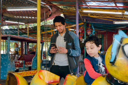 young afro latin father in park using phone without paying attention to his son who is playing alone on machines in mechanical park. family concept, copy space.の写真素材