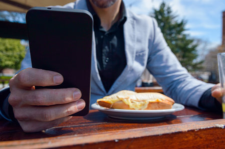 Close-up of unrecognizable Caucasian adult man, sitting at outdoor restaurant, holding phone sending messages with one hand while having sandwich with orange juice for breakfast, copy spaceの写真素材