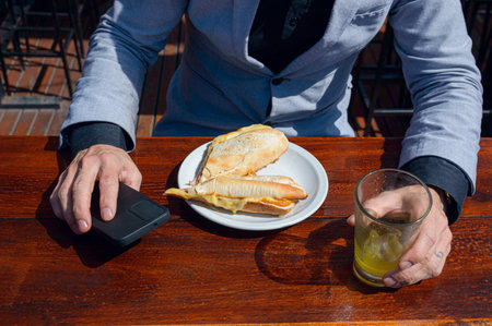 top view of caucasian hands of unrecognizable businessman, sitting outside restaurant, having breakfast of ham and cheese sandwich with orange juice and holding phoneの写真素材