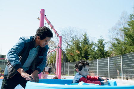 young single afro latino father playing with his son spinning around in plastic bowl, having fun together as family in public park at noon, copy space.の写真素材