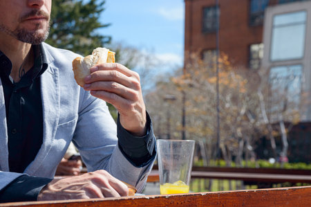 Close-up of unrecognizable adult business man sitting at bar outside restaurant eating sandwich and drinking orange juice.の写真素材