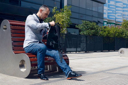 Caucasian adult businessman, sitting outside work buildings looking for something in his backpack, wearing stylish jacket and jean pants, business people concept, copy space.の写真素材