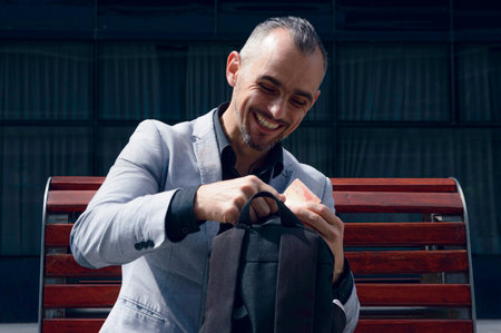 adult businessman, with short hair, beard and wearing stylish gray jacket, is sitting outside,...