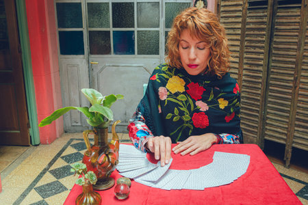 Front view of elegant blonde-haired Latin tarot reader woman sitting at home taking card from tarot...