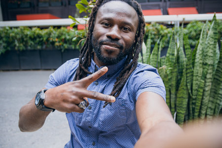Selfie portrait with phone of black man with dreadlocks and beard, sitting looking at camera making...