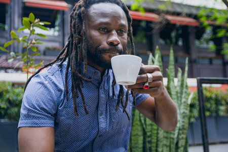 young african man with dreadlocks, beard and casual clothes, sitting outside drinking coffee serious calm looking ahead, holding white cup, copy spaceの写真素材