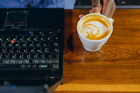 Top view of black male hand holding cup of coffee on cash table, next to black laptop. copy spaceの写真素材