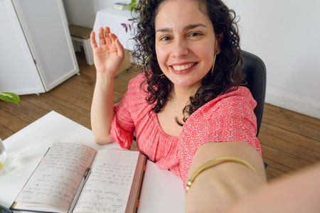Selfie portrait of young Latin Venezuelan woman sitting in her office, happy, waving and smiling...