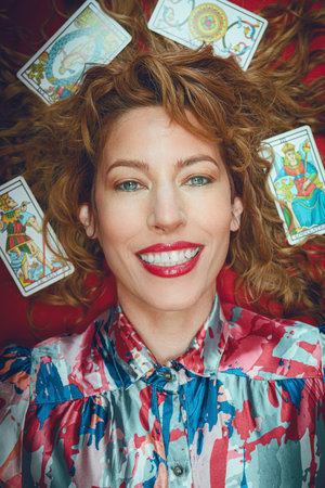 portrait of beautiful happy tarot reader woman smiling looking at camera lying on red table, with...