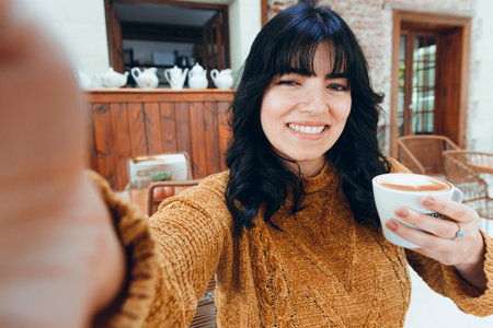 Selfie photo of young Latin Venezuelan woman, in brown, with long black hair, with cup of coffee in her hand, sitting happy and smiling and looking at camera,の写真素材