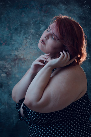 vertical artistic portrait of plus size young latin woman with white skin, with short red hair, standing posing with blue illuminated backgroundの写真素材