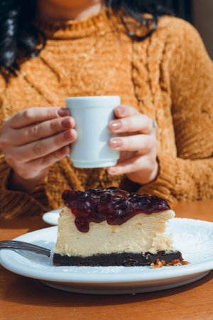 slice of cheesecake with raspberry topping, served on plate on table outdoors, with female hands holding cup of coffee blurred in background, vertical image, copy spaceの写真素材