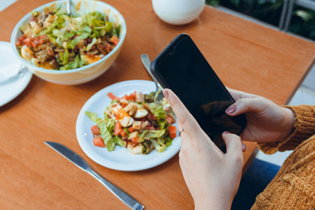 Close-up of caucasian female hands of unrecognizable woman sitting holding phone sending message during lunch, with salad on table in background. copy spaceの写真素材