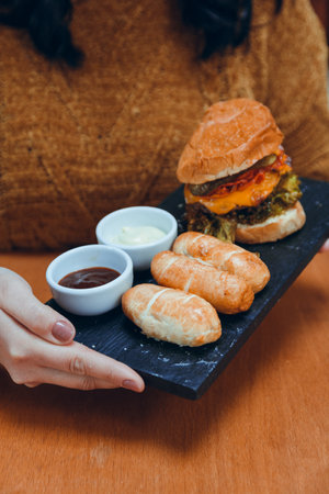 Vertical image of unrecognizable Caucasian woman holding plate of food with Tequenos and served hamburger on stone plate, fast food concept, copy space.の写真素材