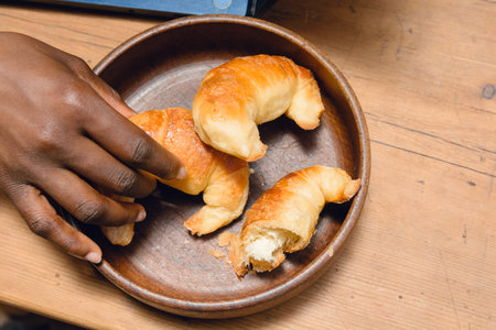 top view of black ethnic female hand indoors, taking sweet croissant from clay bowl on wooden table...