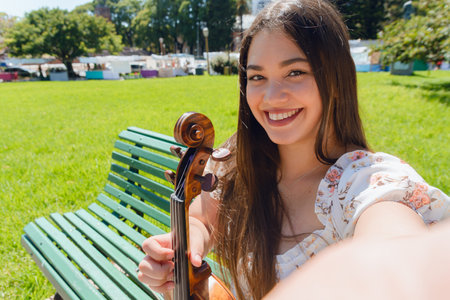 Selfie portrait of young latin busker woman violinist smiling happy on street watching camera holding her violin, standing in plaza in Buenos Aires Argentina.の写真素材