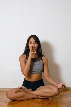 young brunette latina woman at home practicing yoga at home doing breathing exercises and mudras covering her nose with finger. white wall background.の写真素材