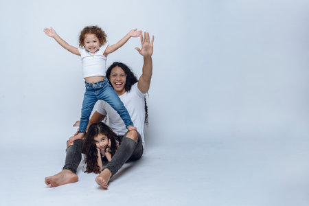 young Latino dad with long hair and glasses, sitting on floor playing with his two young daughters with curly hair, wearing white t-shirt and jean pants, studio photo with white background, copy spaceの写真素材