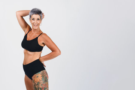 portrait of happy mature hispanic latin woman, with short, conical hair, in underwear with tattoo on her leg, standing laughing happily looking at camera, studio shot, white backgroundの写真素材