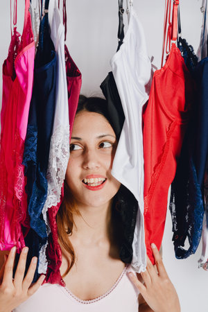 vertical image, close-up portrait of laughing young Latina woman posing inside rack with female underwear, studio photo with white background, photo for lingerie social networks.の写真素材