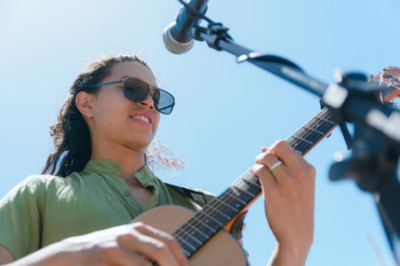 young Latin man with long hair and sunglasses and green shirt, guitarist musician with good attitude standing making rock music with his acoustic guitar outdoors with sky in background.の写真素材