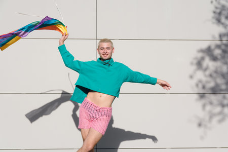 young blond latino man with short hair wearing green sweater and pink shorts dancing with pride flag outdoors, he is very proud for being gay.の写真素材