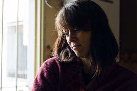 An Adult Latin Woman with short dark hair is sad alone at home standing in a room by the window. She...
