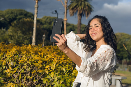 A Young Latin Woman is smiling outdoors taking a picture of herself with her cell phone. She is...